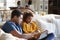 Pre-teen girl sitting on sofa in the living room using tablet computer with her father, mother and toddler sitting at a table in t