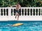 Pre-teens play in the pool by jumping on a small surfboard to keep their balance