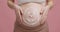 Pregnancy and skin care. Close up of smiley face drawn with cream on big belly, pink studio background, slow motion