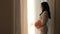 Pregnant beautiful Latin American woman standing looking out at window and caressing her stomach