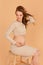 Pregnant beautiy girl in a long beige clothes on a beige background in the studio. Fashionable style for pregnant women. Studio