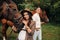 A pregnant girl in a hat and her husband in white clothes stand next to horses in the forest in nature.Stylish pregnant woman with