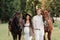 A pregnant girl in a hat and her husband in white clothes stand next to horses in the forest in nature.Stylish pregnant woman with