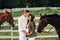 A pregnant girl in a hat and her husband in white clothes stand next to the horses near the horse corral.Stylish pregnant woman