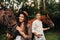 A pregnant girl in a hat and a man in white clothes stand next to horses in the forest in nature.Stylish pregnant woman with her