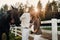 A pregnant girl in a hat and a man in white clothes stand next to horses near a white fence.Stylish pregnant woman with a man with