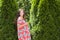 Pregnant girl near beautiful greenery. Beautiful pregnant woman in dress and touching belly stands near thuja. Future mother in a