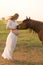 A pregnant girl in white communicates with a horse on a green meadow at sunset. Therapy and relaxation for pregnant women.