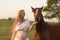 A pregnant girl in white communicates with a horse on a green meadow at sunset. Therapy and relaxation for pregnant women.