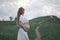 A pregnant girl in white is waiting for her beloved stand by a hill with a beaten track. Rural natural landscape