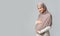 Pregnant muslim woman embracing her belly, posing over grey background