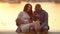 Pregnant parents sitting on pier of lake at sunset and embracing stomach.