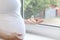 A pregnant, sick second-trimester woman in a white T-shirt is holding a glass jar of pills near a dirty window. Copy space -