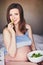 Pregnant smiling girl in T-shirt sit on bed and eat salad with green and red leaves. Young woman keep diet during pregnancy. Healt