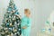 Pregnant smiling woman wearing blue dress hugging belly and decorating Christmas tree.