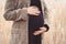 Pregnant unrecognizable woman in black dress standing in reeds and holding her belly.