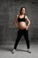 Pregnant woman with belly, in black sport pants and top, pink sneakers. Hands on hips. Posing on gray background. Full-length