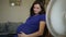 A pregnant woman in a blue dress is sitting on a swivel chair in front of a fan.