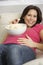 Pregnant Woman Eating Bowl Of Popcorn Sitting On Sofa At Home