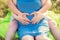 Pregnant woman and her husband sitting in the park and holding their hands in a heart shape on her baby bump. Close up