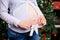 A pregnant woman holds in her hands children`s socks, booties on the background of a Christmas tree. Dressed in a shirt and junks