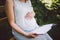Pregnant woman reads a book on a bench and holds her hand on her belly in the summer