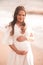 Pregnant woman showing heart shape with fingers wearing white dress over sea background. Motherhood. Maternity.