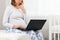 A pregnant woman sits on the bed with a thoughtful face and uses a laptop. Concept of modern technologies and freelancing. Close