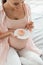 Pregnant woman sitting on bed and holding saucer with cupcake