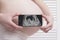 Pregnant woman in underwear holding and showing mobile phone with fetal ultrasound photo on white wooden screen background. Close-