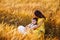 A pregnant woman in a yellow dress with daughter in white sundresses sit hugging in a wheat field