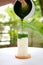 Preparing iced matcha green tea with milk in a square glass over garden background