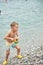Preschooler boy carries toy watering can on pebble beach