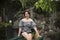 A pretty asian woman wearing summer attire sitting on a faux bamboo bridge at a lush stone garden landscape