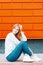 Pretty attractive european stylish young woman in blue jeans sneakers in a sweater is sitting on the sidewalk