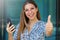 Pretty beautiful young woman with thumb up holds smart phone and looks at camera