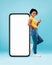 Pretty black lady standing near big smartphone with blank screen, gesturing thumb up on blue background, mockup