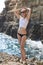 Pretty blonde in white tank top and black bikini bottom posing on background of sea edge