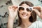 Pretty brunette girl smiling, trying on sunglasses in optical shop. Charming feminine woman in sunglasses store fooling