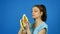 Pretty Brunette Young Woman Holding in Hand Yellow Banana and Then Eating it over Bright Blue Background. Tropical