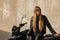 Pretty Caucasian lady wearing a leather jacket with an all-black outfit next to her motorbike