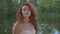 Pretty caucasian woman with red hair in beige dress standing on the bank of the lake and looking up and around. Redhead