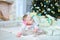 Pretty female baby lying on floor near twinkling Christmas tree and presents.