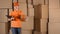 Pretty female courier in orange uniform delivering a parcel against cardboard boxes stacks backround. 4K studio video