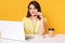 Pretty female sits at white desk with opened laptop computer, writes email, uses high speed internet, poses isolated over yellow