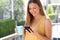 Pretty girl having breakfast on balcony in sunny morning. She holds a mug and reading friends messages on mobile phone