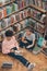 Pretty girl and her classmate sitting on the floor by large bookshelf in library