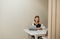 Pretty girl sitting in apartment at desk with laptop and smartphone in her hands, working and looking at camera with serious face