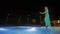The pretty girl walking on the edge of outdoor open pool on roof of hotel. Night view of city lights. Girl in swimwear