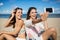 Pretty girls sitting on beach taking selfie laughing
